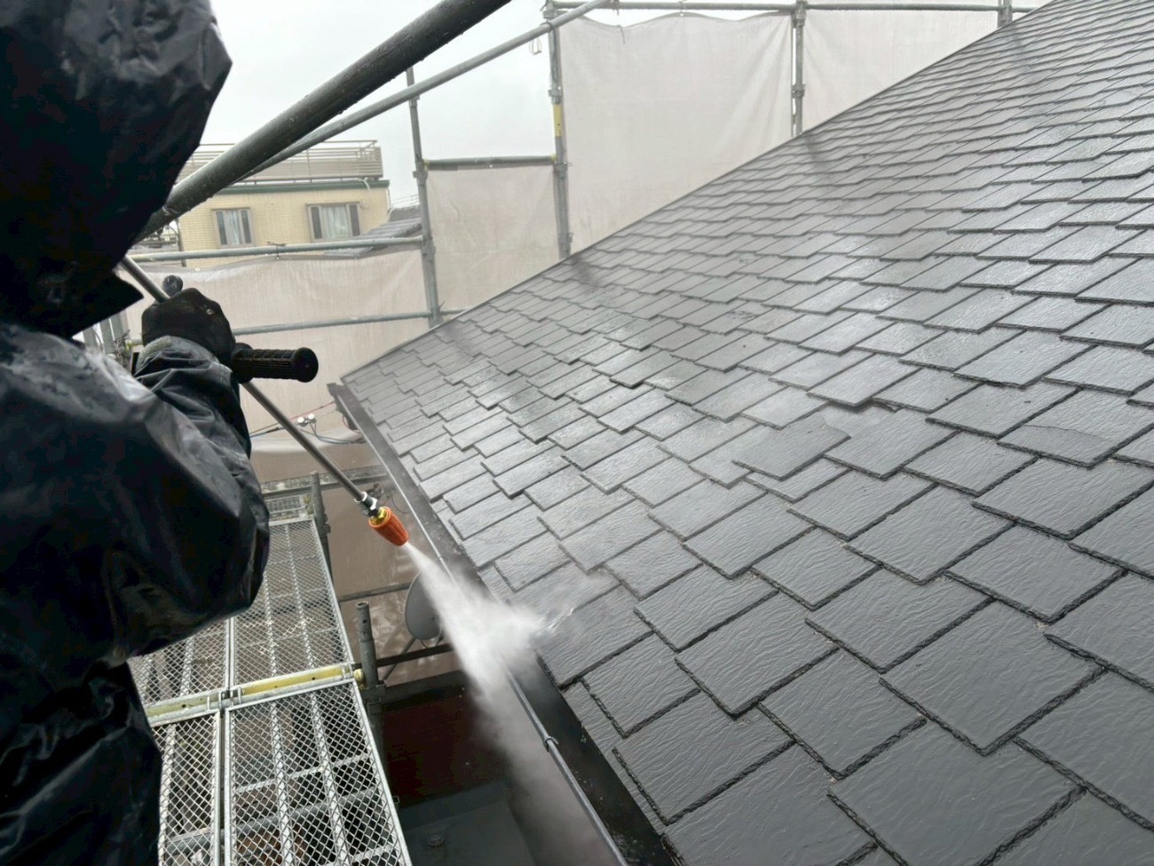 足場から屋根の雨樋内部を高圧洗浄する職人