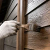 白い作業手袋を着けた手が、刷毛で木目のある外壁板に茶色の塗料を塗っている