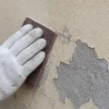 白い作業用手袋を着けた手が、塗膜の剥がれた壁を紙やすりでこすっている