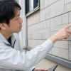 作業着姿の男性が建物の灰色の外壁に顔を近づけ、タイルの目地を指で確認している