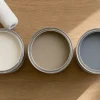 木目の台に白、ベージュ、グレーの塗料缶が並び、ローラーと刷毛が置かれている