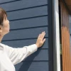 白い服を着た女性が、屋外で住宅の紺色の横張り外壁に右手で触れて見上げている