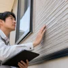 白い作業着の男性がタブレットを持ち、住宅の窓横にある外壁を手で触れて確認している