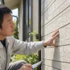 作業着の男性が屋外で建物の外壁に手を当て、クリップボードを持ちながら表面を確認している