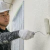 白いヘルメットと手袋を着けた作業員が、ローラーで建物の外壁を白く塗っている